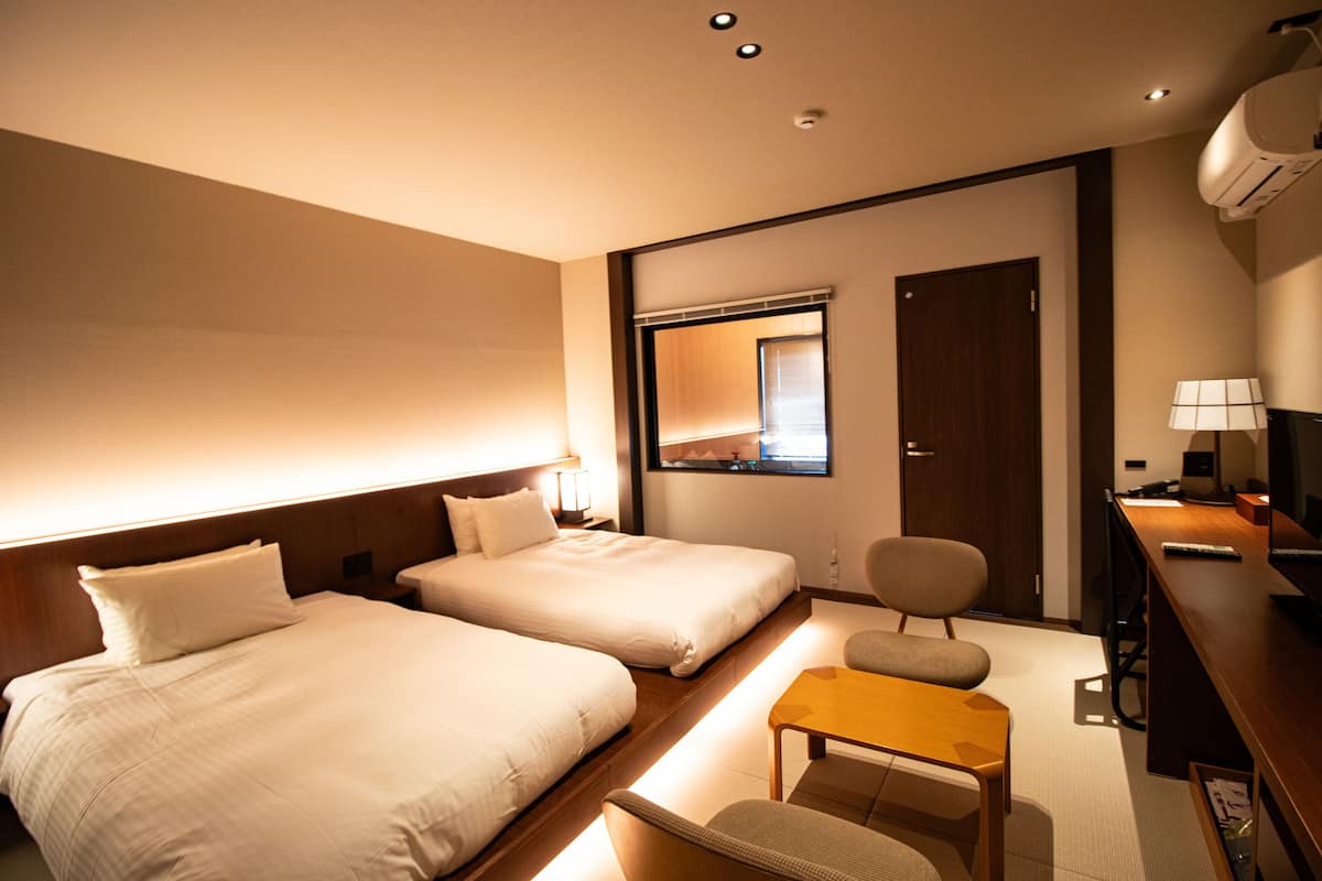 A hotel room with two beds, a desk, a chair, a small table, a mirror, and a door.