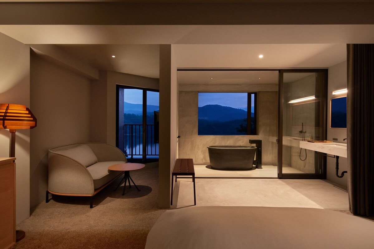 A modern hotel room with a bed, a seating area with a table, a bathtub, and a large window offering a mountain view.