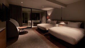 Ett hotellrum med två sängar, ett skrivbord, en stol, en glasdörr som leder ut till en balkong och en vägglampa.
