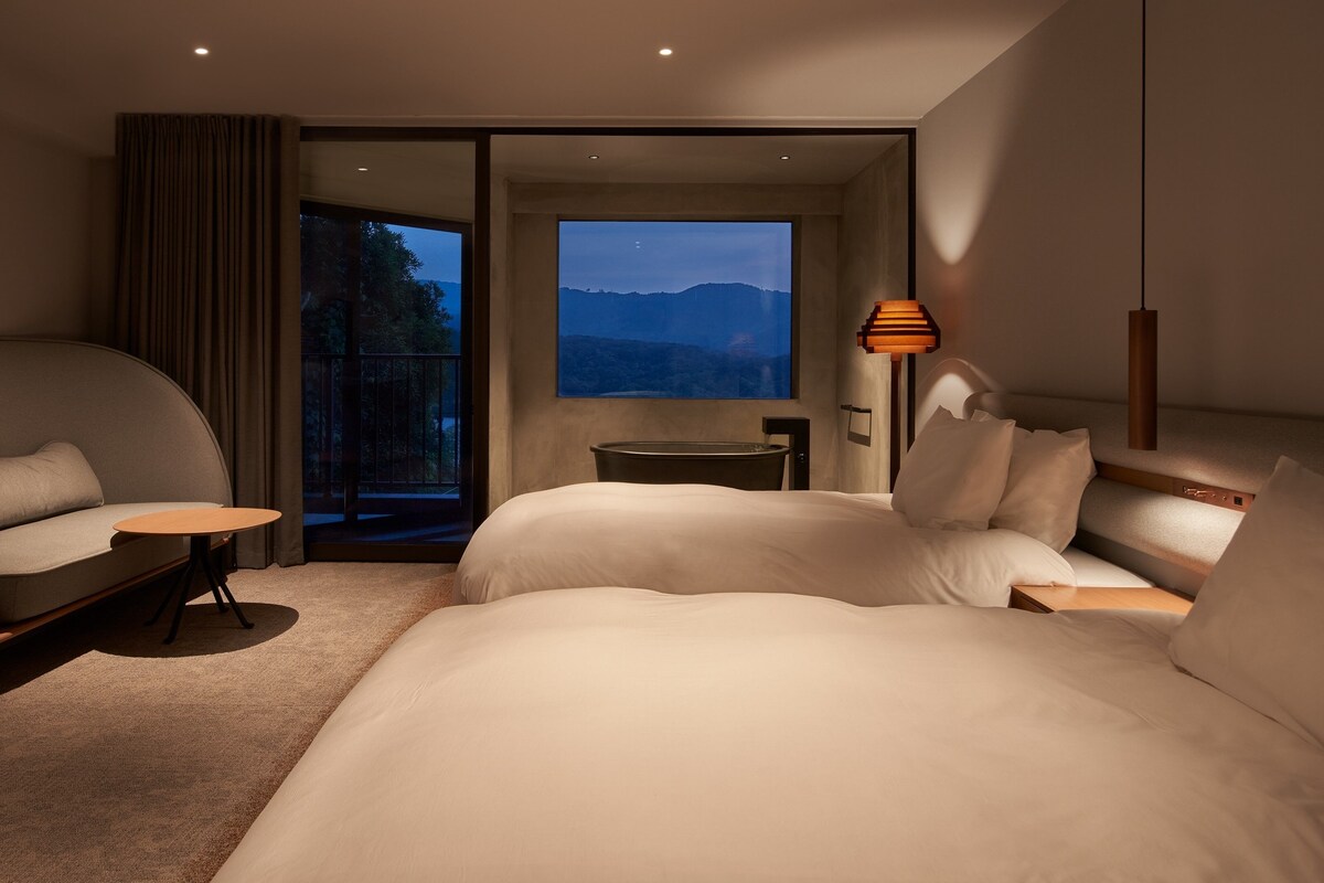 A modern hotel room with a large bed, a sofa, a small table, and a view of mountains.
