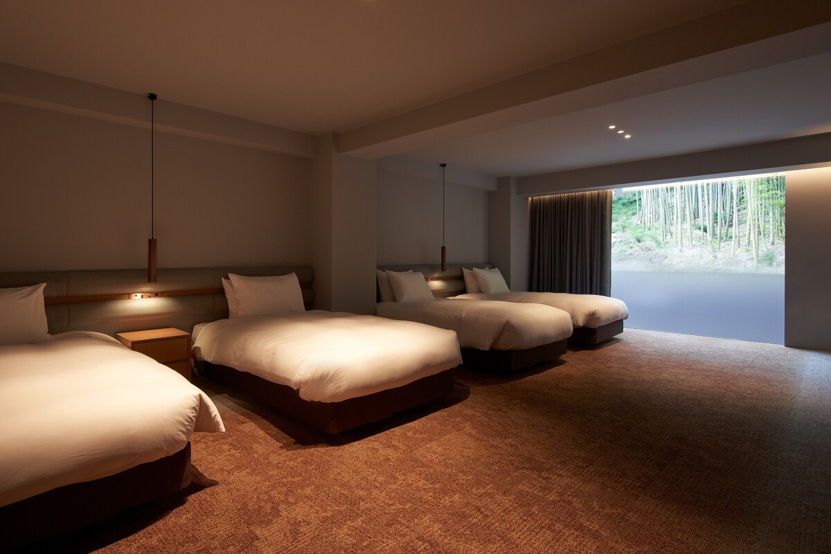 A modern hotel room with three beds, a large window with a view of trees, and a carpeted floor.
