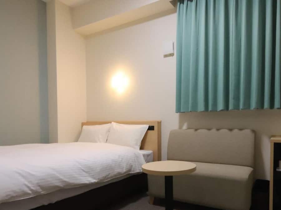 Una habitación de hotel con una cama, una mesita redonda pequeña, un sofá y una lámpara fijada a la pared.