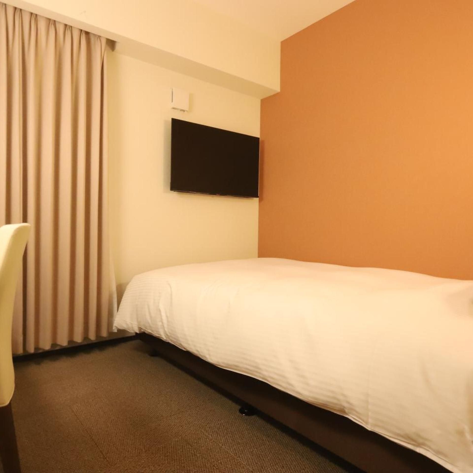 A hotel room with a bed, a chair, a flat-screen TV mounted on the wall, and a carpeted floor.