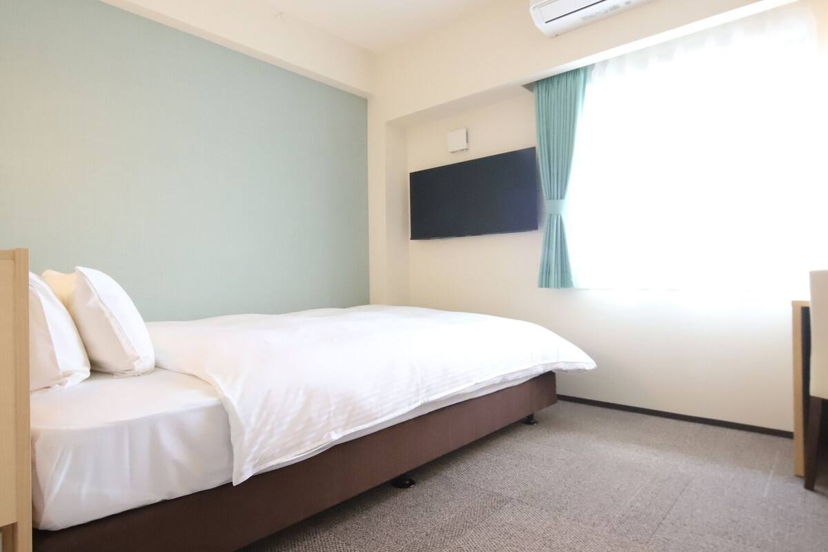 Ein Hotelzimmer mit einem Bett, einem an der Wand befestigten Flachbildfernseher, einem Fenster mit Vorhängen und einem kleinen Schreibtisch mit Stuhl.