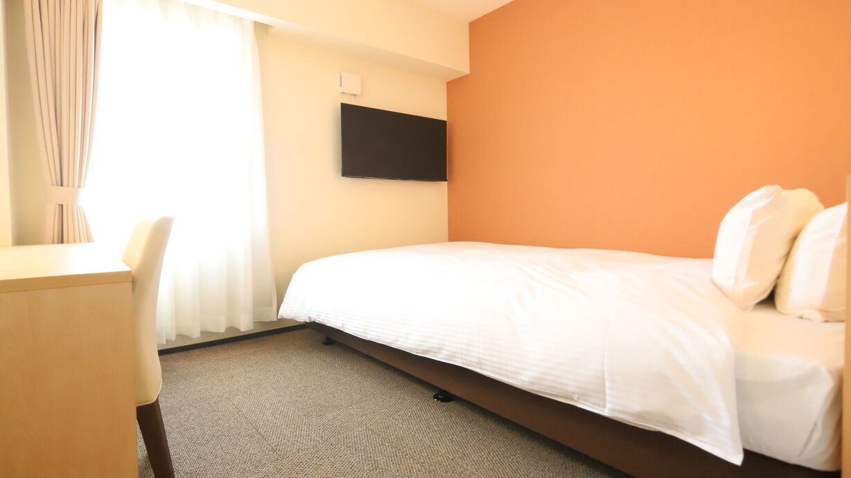 Habitación de hotel con cama, escritorio, silla, televisor de pantalla plana montado en la pared y ventana con cortinas.