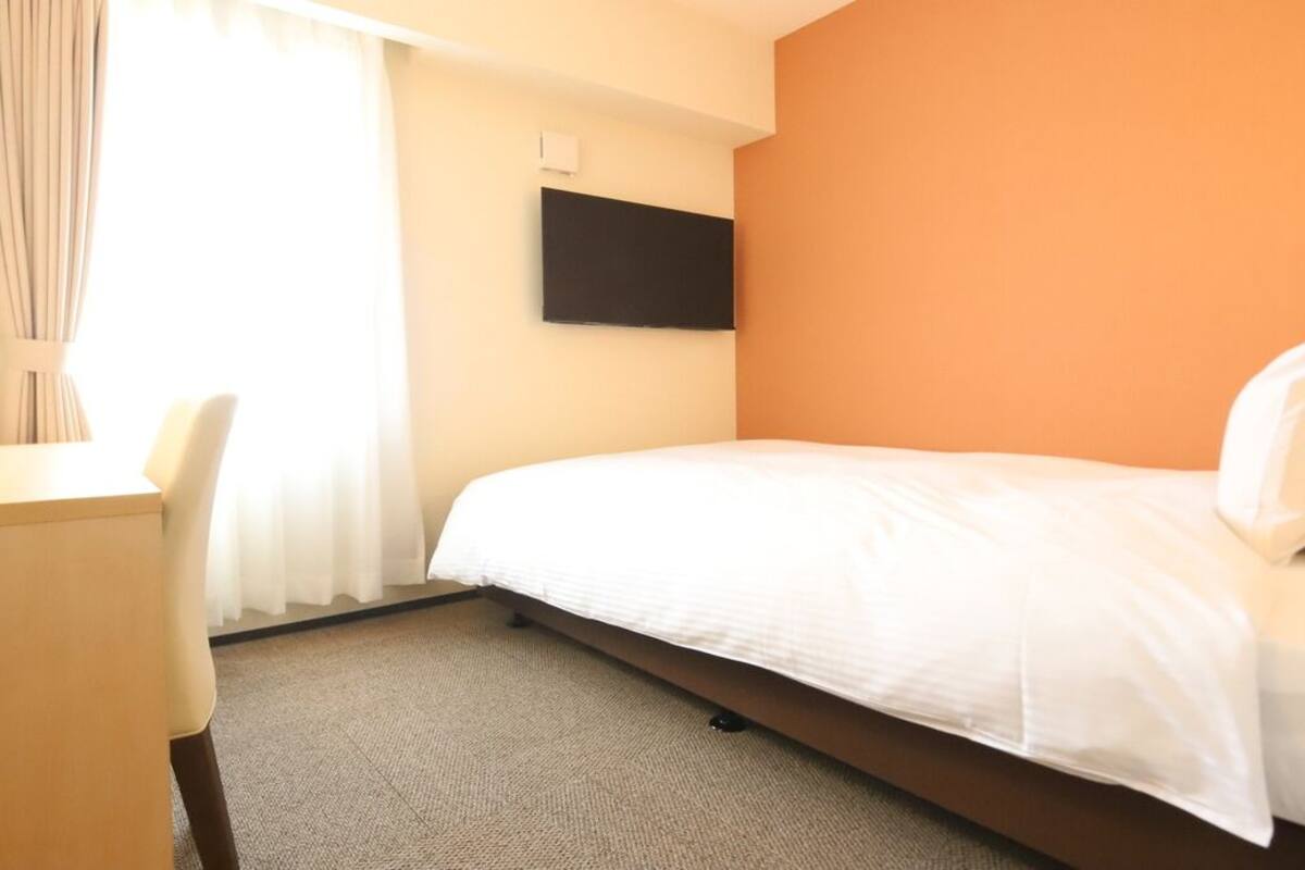 Ein Hotelzimmer mit Bett, Schreibtisch, Stuhl, einem an der Wand befestigten Flachbildfernseher und einem Fenster mit Vorhängen.