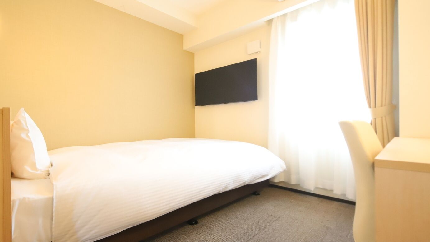 Una habitación de hotel con una cama individual, un televisor de pantalla plana montado en la pared, un escritorio con una silla y una ventana con cortinas.