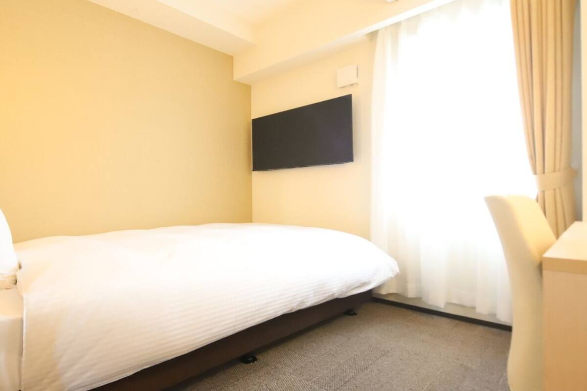 Ein Hotelzimmer mit einem Einzelbett, einem flachbildfernseher an der Wand, einem Schreibtisch mit Stuhl und einem fenster mit Vorhängen.