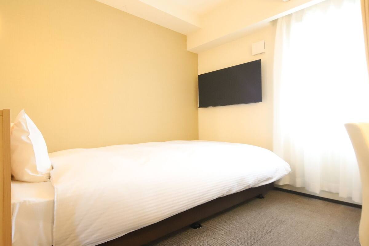 Ein Einzelbett mit Kopfstütze, ein Nachttisch, ein flachbildfernseher an der Wand und ein Fenster mit Vorhängen.