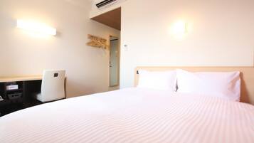 A hotel room with a bed, a desk, a chair, an air conditioning unit, and a wall-mounted light fixture.