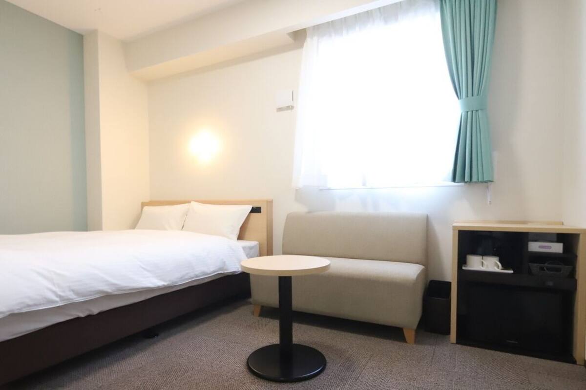 Ein Hotelzimmer mit Bett, kleinem runden Tisch, Sofa, Nachttisch und einem Fenster mit Vorhängen.