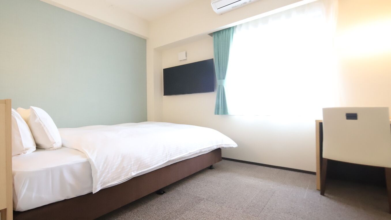 Una habitación con una cama, un televisor de pantalla plana en la pared, una ventana con cortinas y un escritorio pequeño.