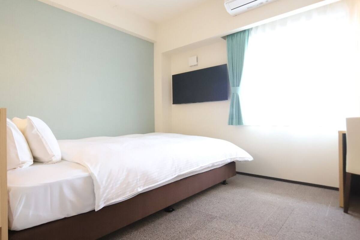 Ein Zimmer mit einem Bett, einem flachbildfernseher an der Wand, einem Fenster mit Vorhängen und einem kleinen Schreibtisch.