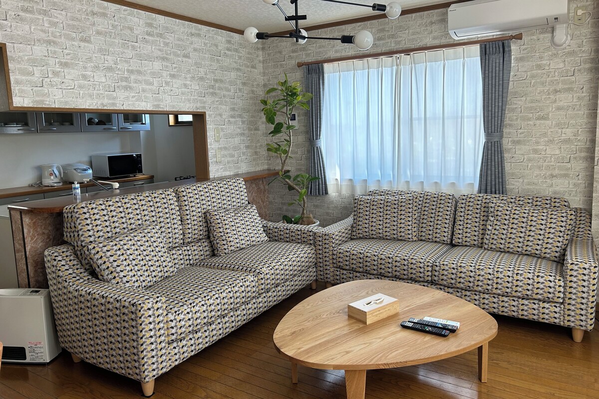 A living room with a sectional sofa, a coffee table, a potted plant, and a kitchen area in the background.
