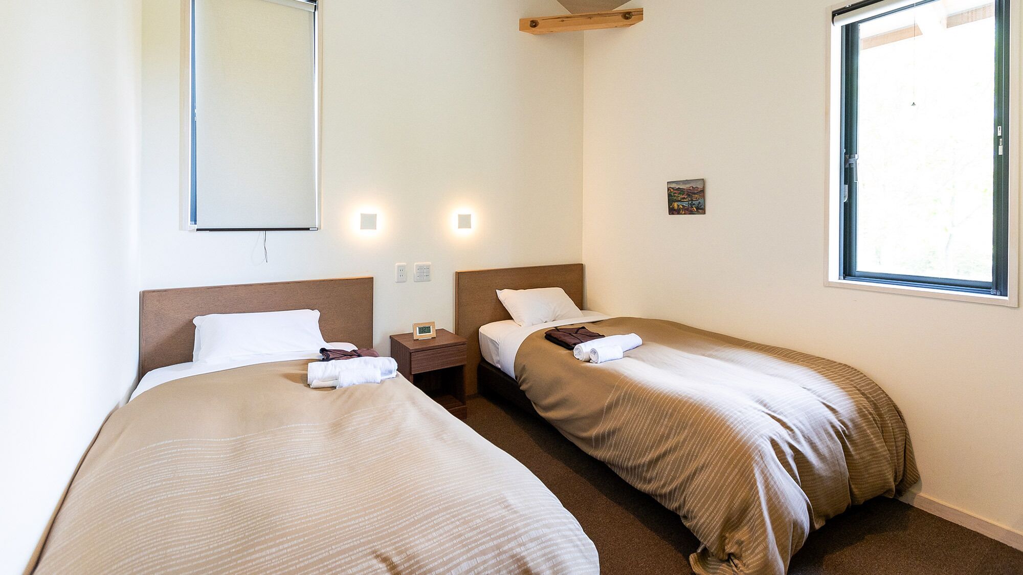 Habitación de hotel con dos camas, una mesita de noche con un reloj, un pequeño cuadro en la pared y una ventana.