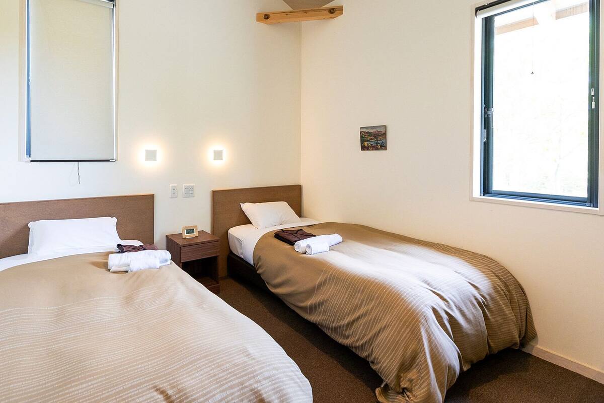 A hotel room with two beds, a bedside table with a clock, a small painting on the wall, and a window.