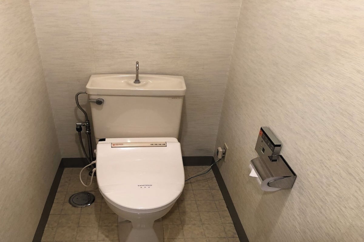 Un piccolo WC bianco con maniglia per lo sciacquone e un accessorio bidet, un porta carta igienica a parete e uno scarico a pavimento.