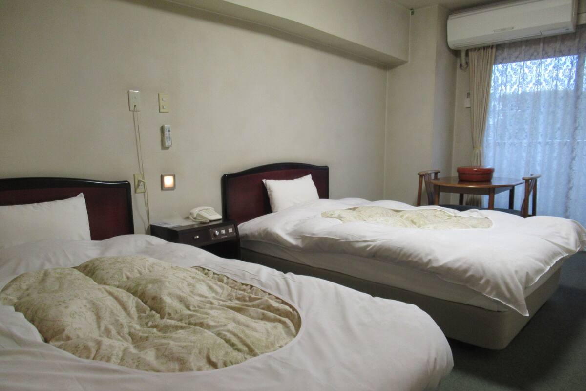 Een hotelkamer met twee bedden, een nachtkastje met een telefoon, een klein tafeltje met een schaal en een airconditioner.