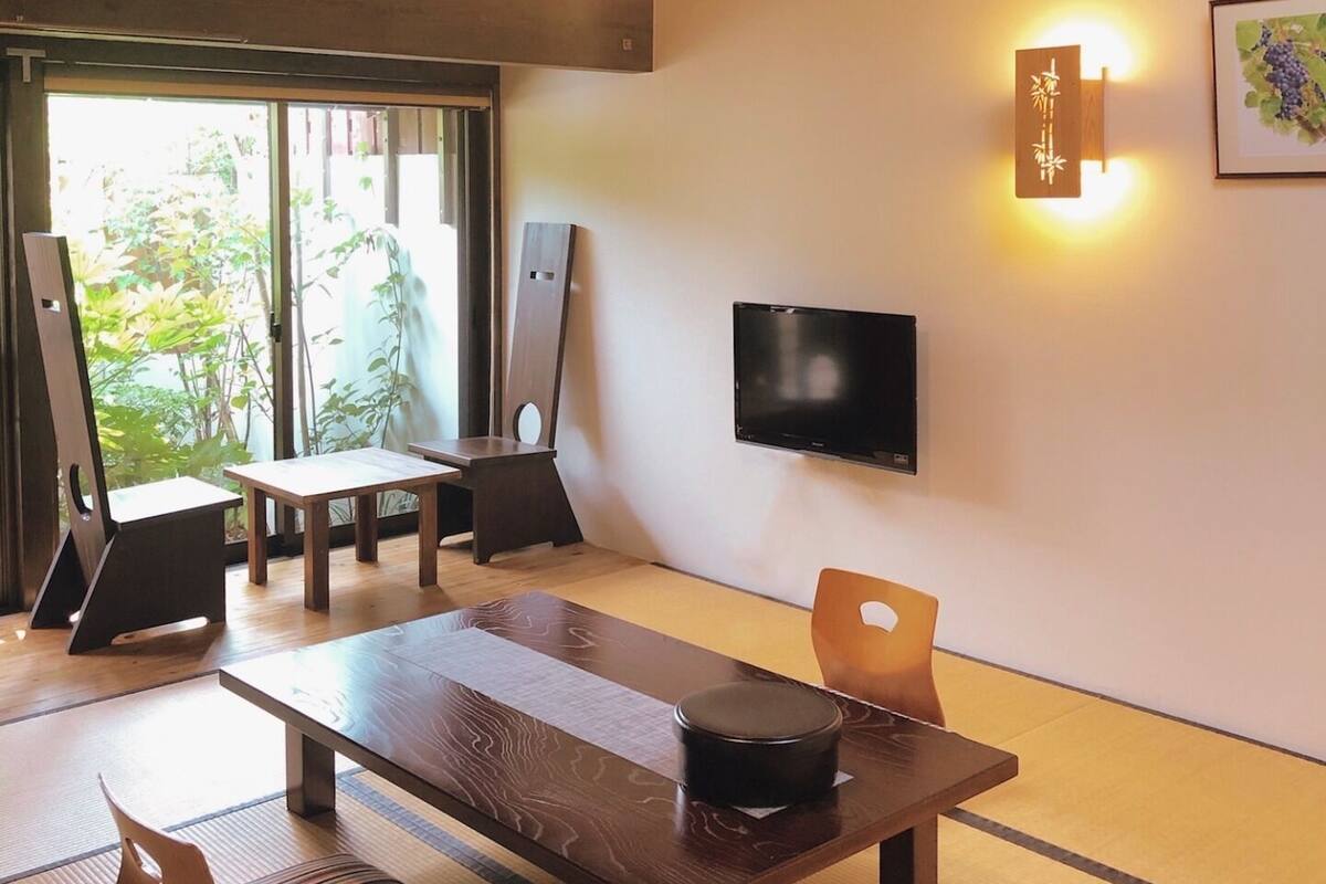 Una habitación tradicional japonesa con suelo de tatami, una mesa baja, una silla, un televisor y una lámpara fijada a la pared.