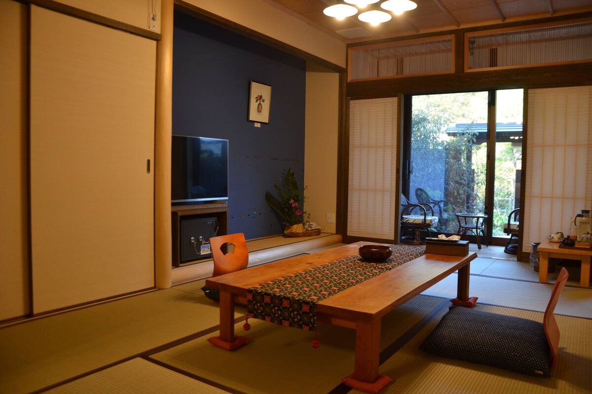 Una habitación japonesa tradicional con suelo de tatami, una mesa baja con mantel tejido, un televisor y una puerta corredera que da a un espacio exterior.