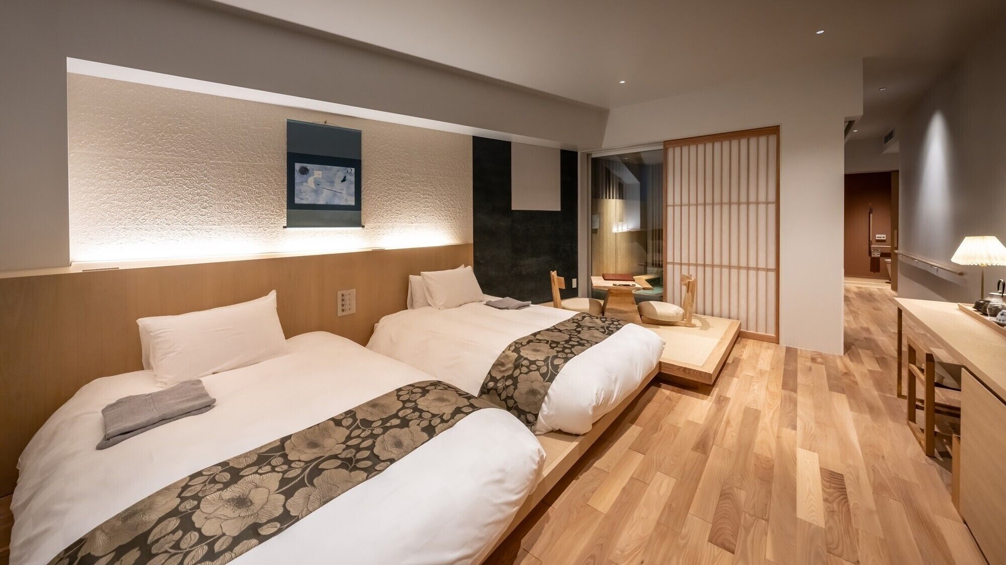 Habitació d'hotel amb dos llits, un panell de fusta al capçal, un escriptori petit i una porta corredissa que condueix a un bany.