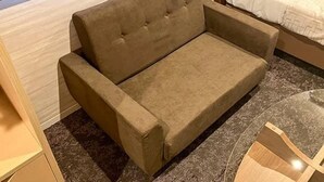 A brown sofa in a hotel room with a wooden floor and a bed with white bedding.