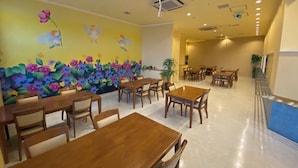 A dining area with wooden tables and chairs, a colorful mural with flowers and angels, and ceiling lights.