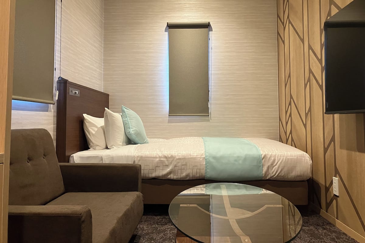 Une chambre d’hôtel de taille réduite, comprenant un lit, un petit canapé, une table basse en verre, un téléviseur et une fenêtre avec des stores.