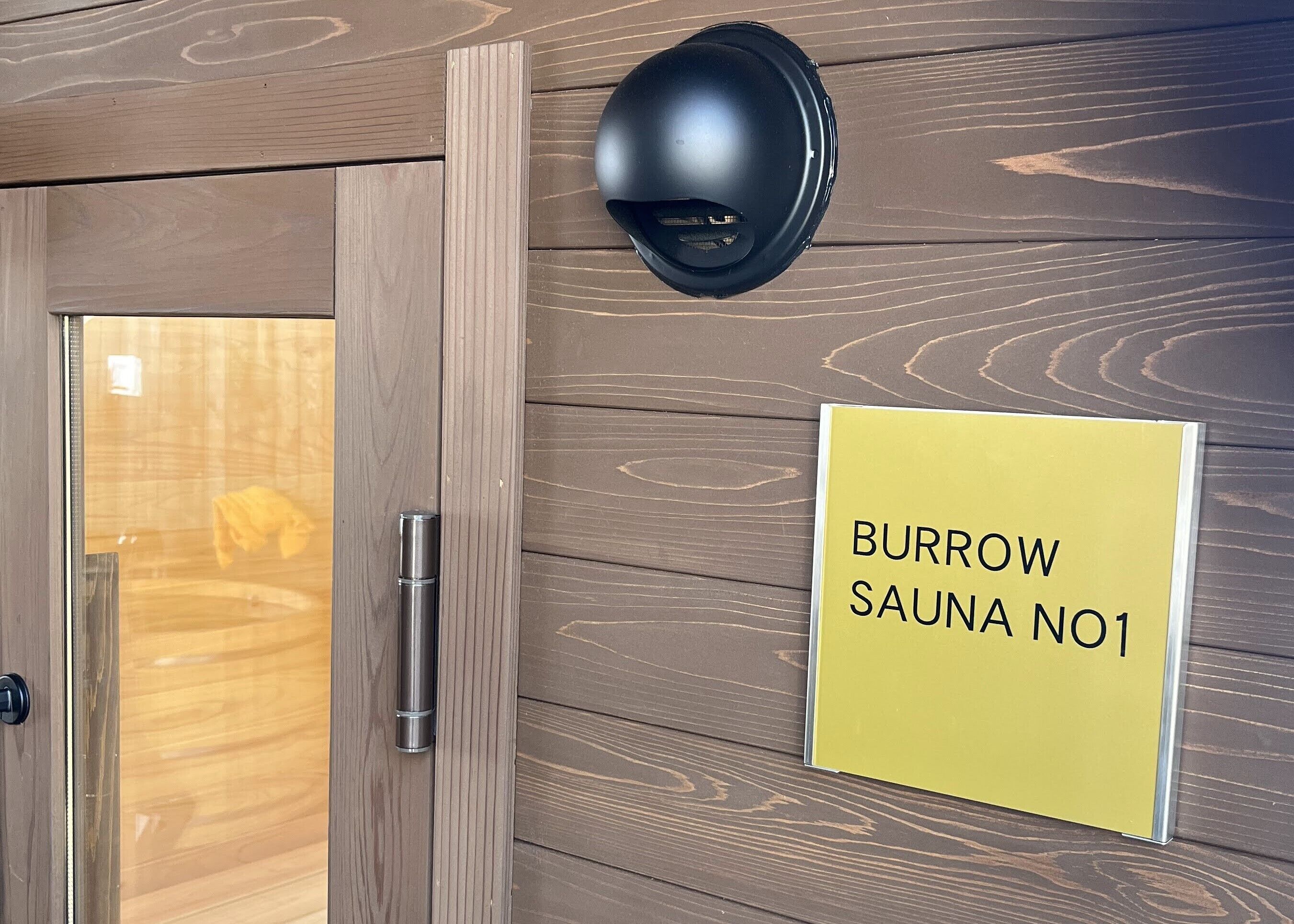 Pared de madera con un letrero que dice 'BURROW SAUNA NO1' y una cámara de seguridad negra sobre una puerta.