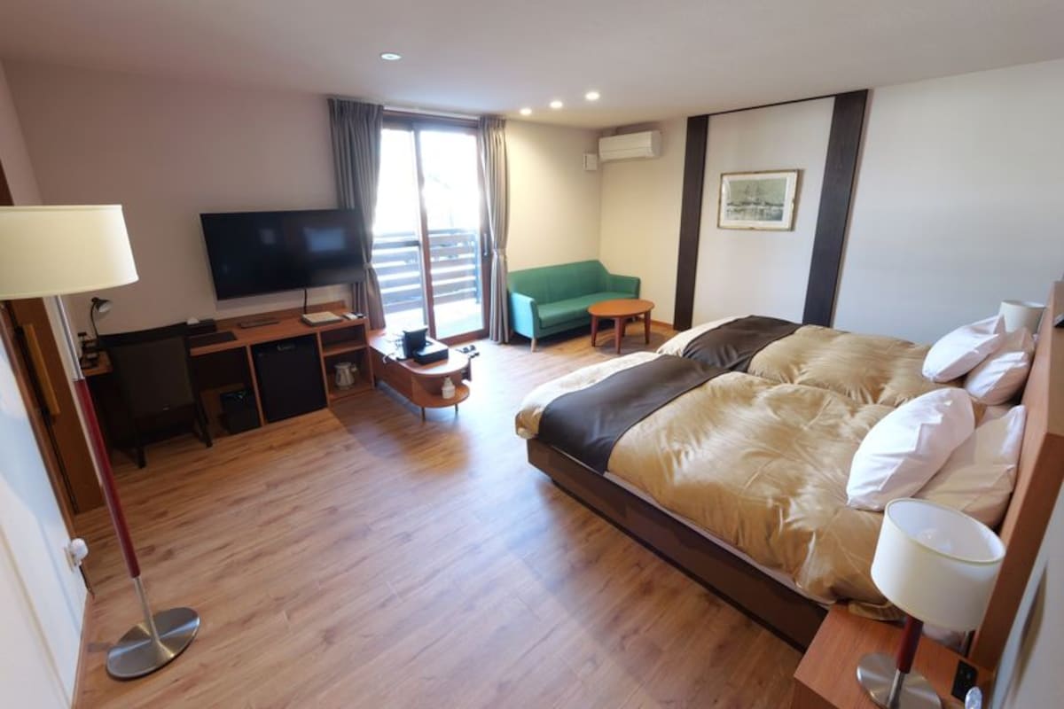 A hotel room with a bed, a desk with a TV, a green sofa, and a small table.