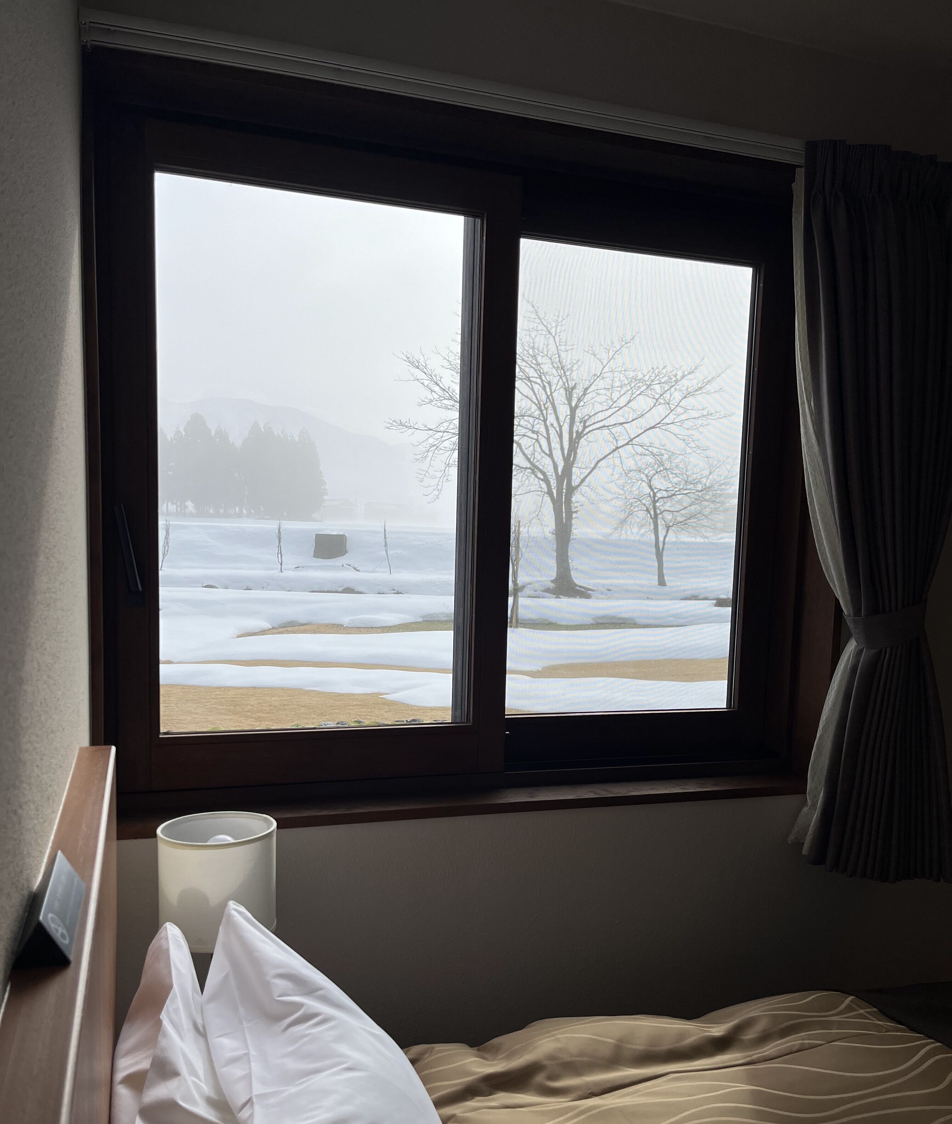 Una habitación con cama, una ventana con cortinas, una mesita de noche con lámpara y vistas a un paisaje nevado.