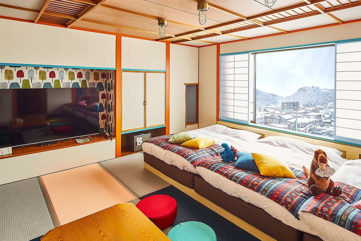 Ein traditioneller japanischer Raum mit Tatami-Böden, einem Bett mit farbenfroher Bettwäsche, einem Fernseher und Blick auf eine verschneite Landschaft.