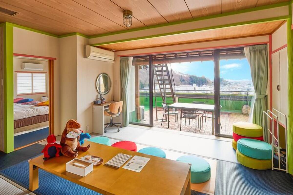 A room with a wooden ceiling, a bed with a window, a desk with a chair, a table with colorful cushions, and a view of a snowy landscape through large glass doors.