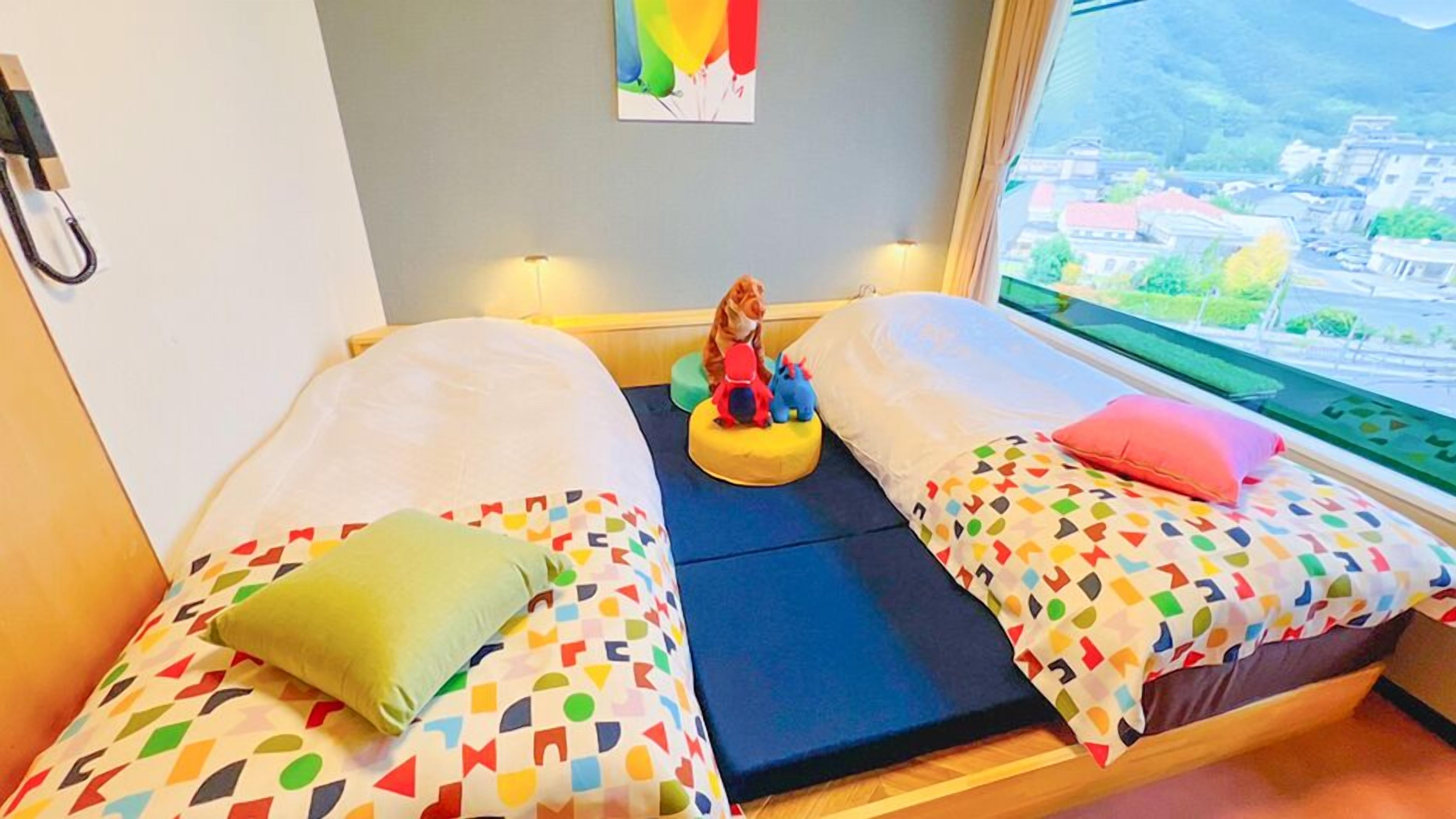 Ein Zimmer mit zwei Betten, bunte geometrische Bettwäsche, einem kleinen Tisch mit Spielzeug, einem Bild an der Wand und einem Fenster mit Blick auf die Stadt.
