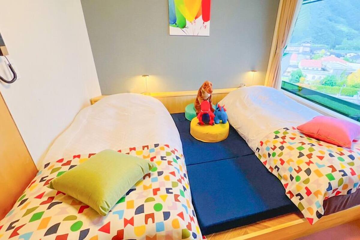 A room with two beds, colorful geometric-patterned bedding, a small table with toys, a painting on the wall, and a window with a city view.