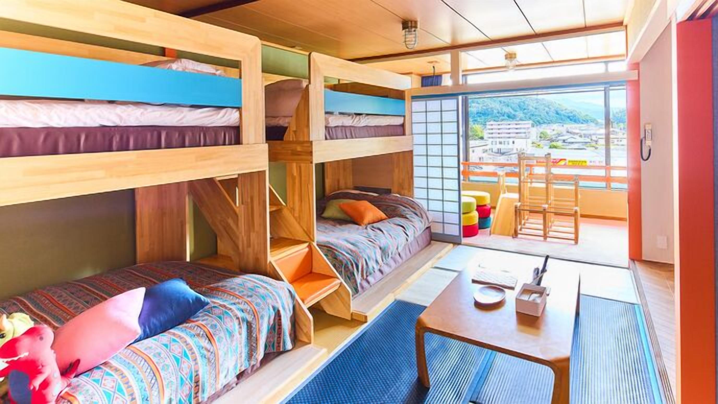 Ein Schlafsaalzimmer mit Etagenbetten, einem kleinen Tisch und einem Stuhl sowie Blick auf eine Stadt durch ein großes Fenster.