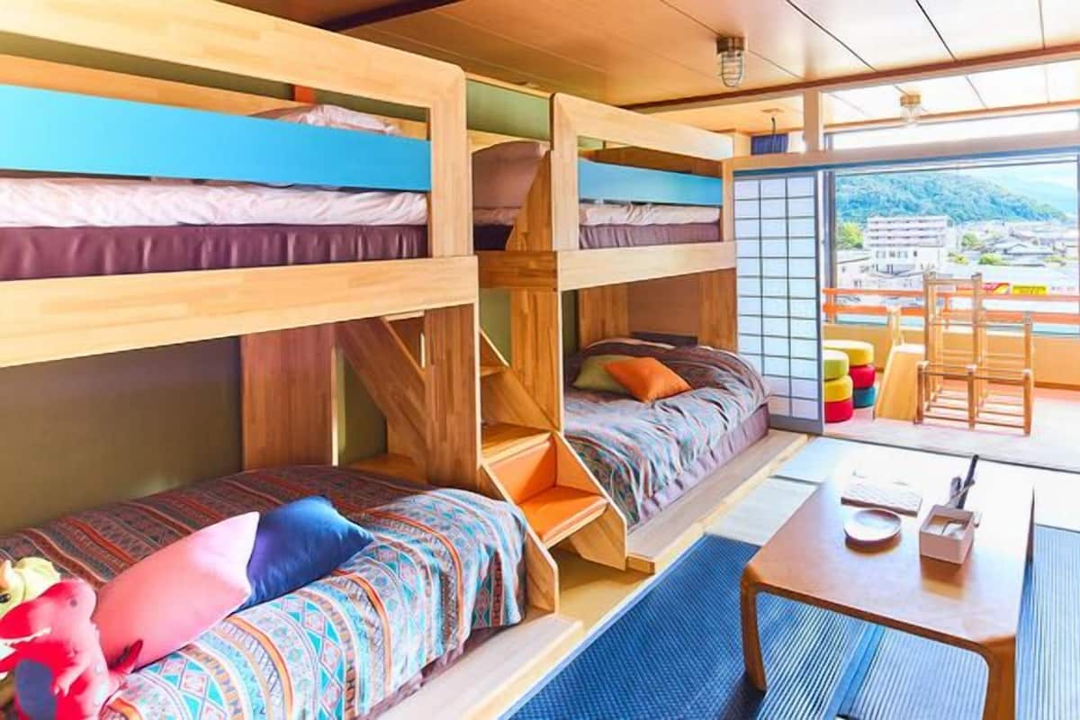 Ein Schlafsaalzimmer mit Etagenbetten, einem kleinen Tisch und einem Stuhl sowie Blick auf eine Stadt durch ein großes Fenster.