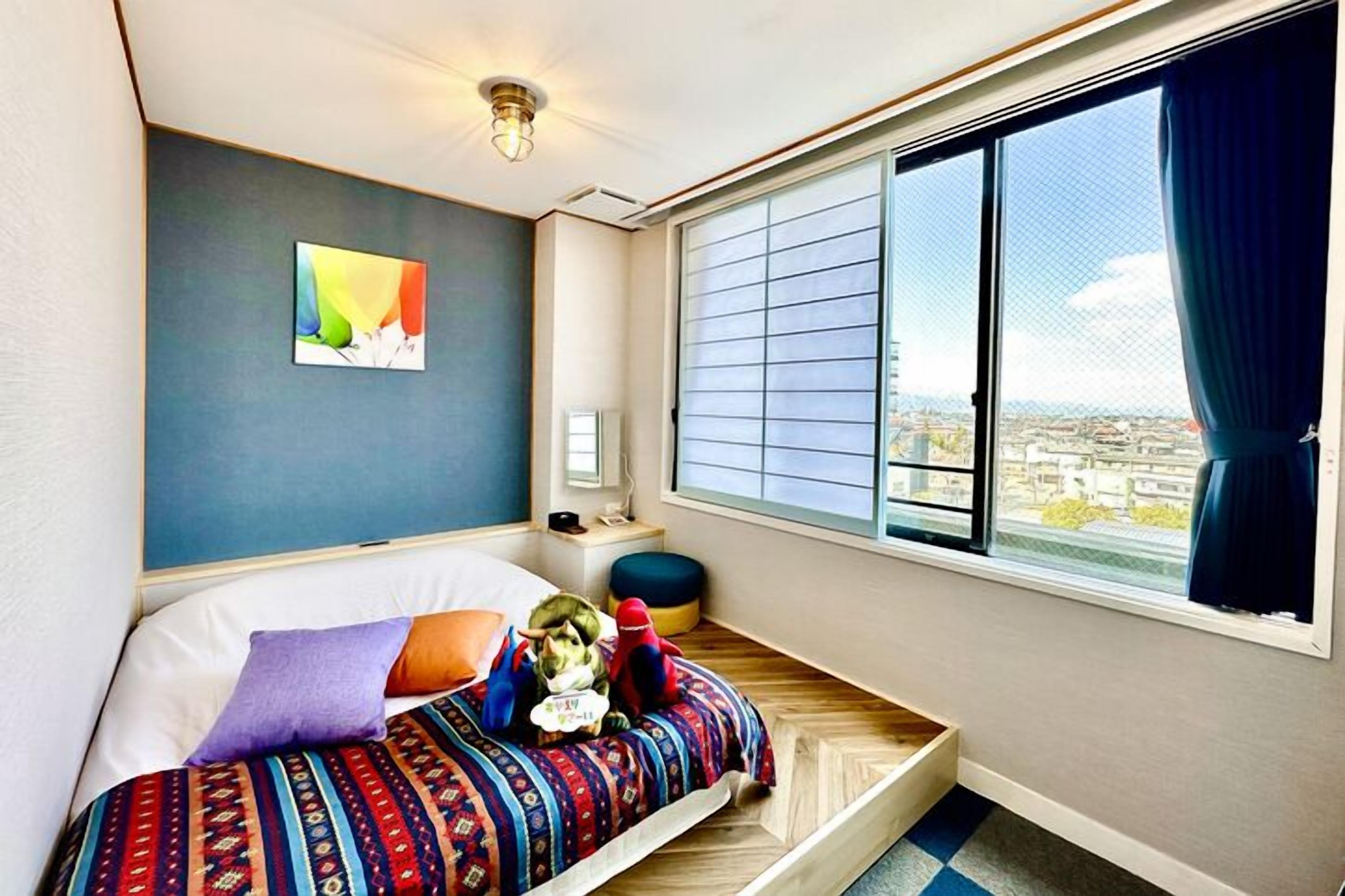 Ein Schlafzimmer mit Bett, einem Fenster mit Jalousien, einer Wandleuchte und Blick auf die Stadt.