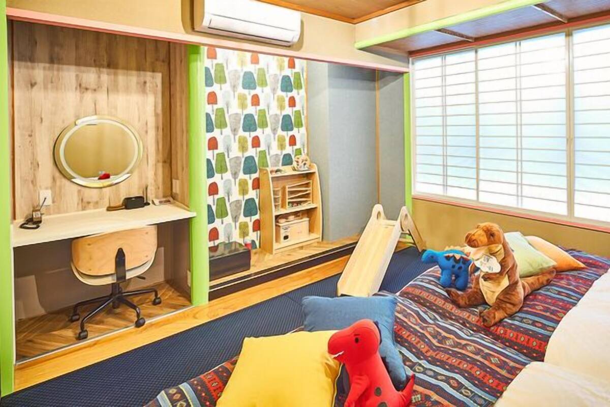 A bedroom with a bed, a desk with a chair, a window with blinds, and a shelf with toys.