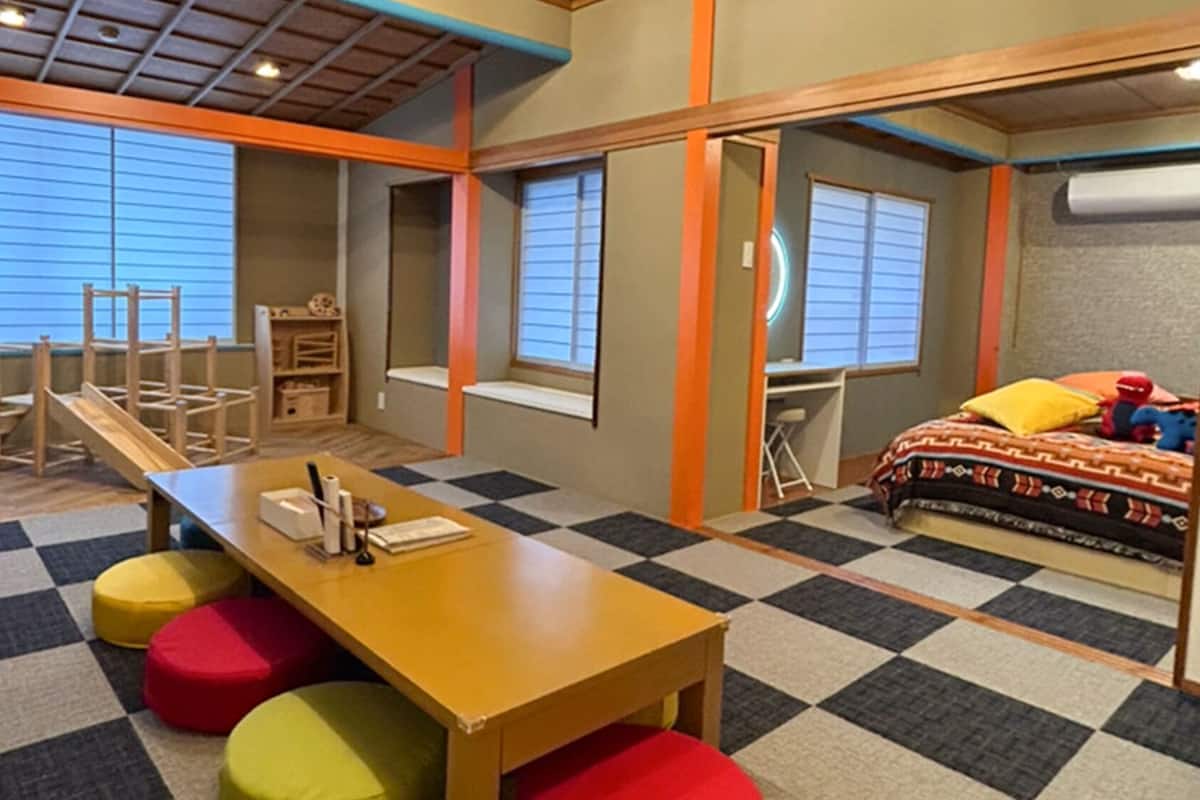 Ein Zimmer mit Schachbrettfußboden, einem Holztisch, bunten Hocker, einem Bett mit gemusterten Textilien und einem Fenster mit Jalousien.