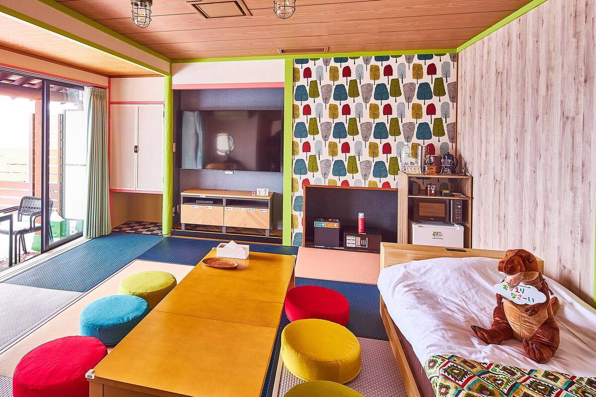 A room with a bed, a table, colorful cushions, a TV, and a patterned wall.