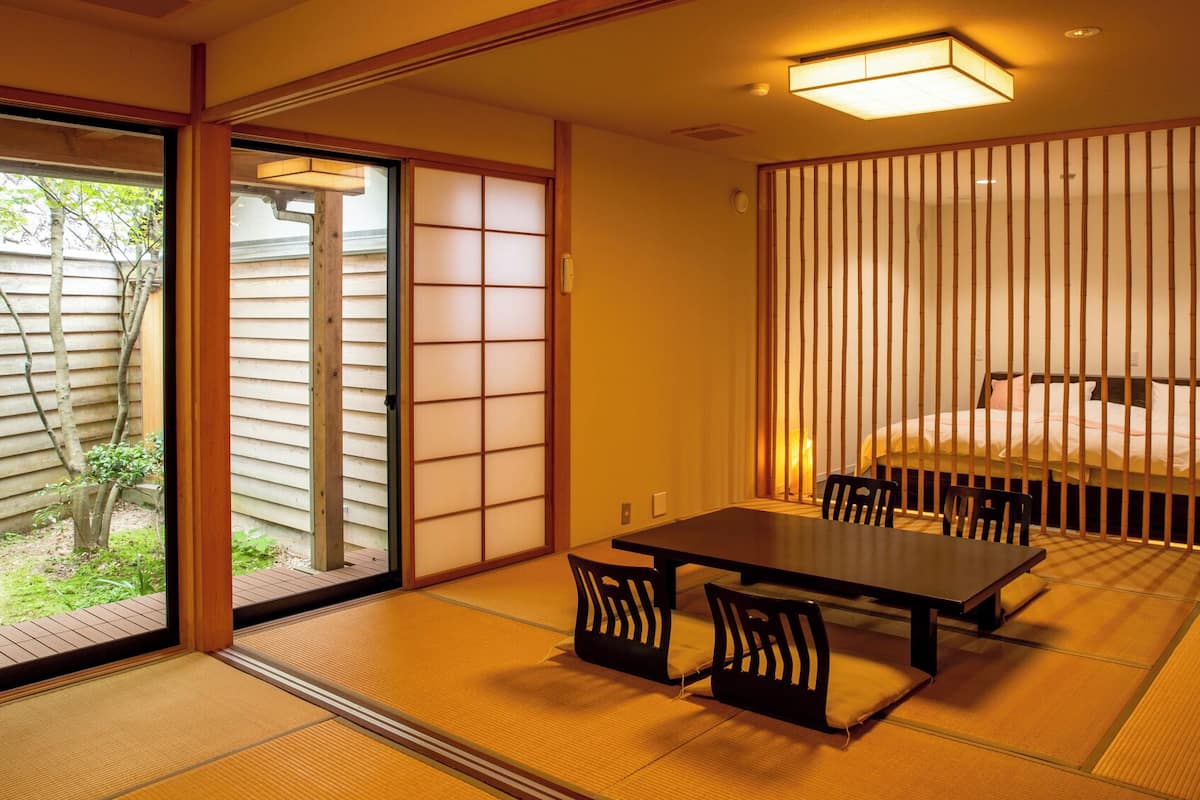 A traditional Japanese room with tatami flooring, a low table with chairs, a sliding door, and a bed with a headboard.