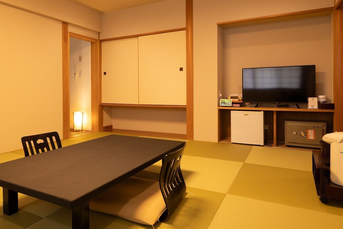 A room with tatami flooring, a low table, chairs, a television, a refrigerator, and a safe.
