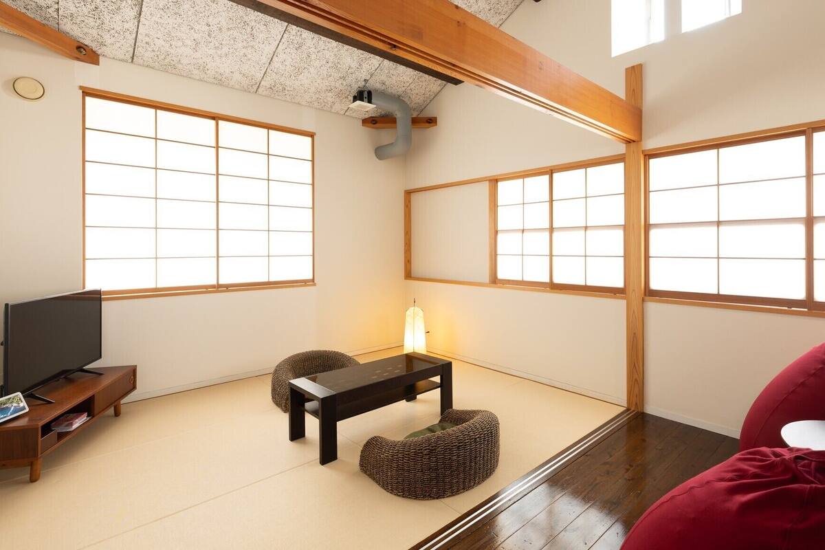 A traditional Japanese room with tatami flooring, sliding shoji doors, a television, a small table, and a red bean bag.