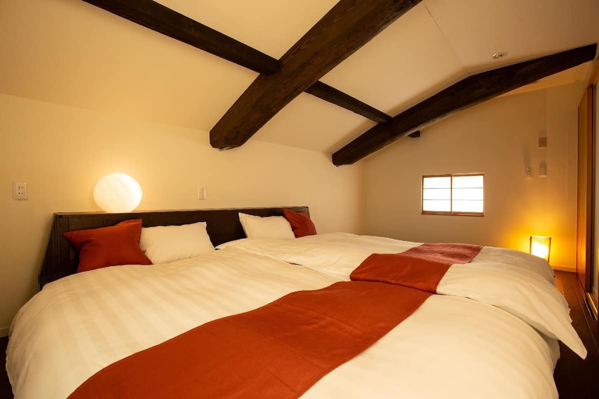 A bedroom with a large bed, wooden beams on the ceiling, a window, and a wall-mounted light.
