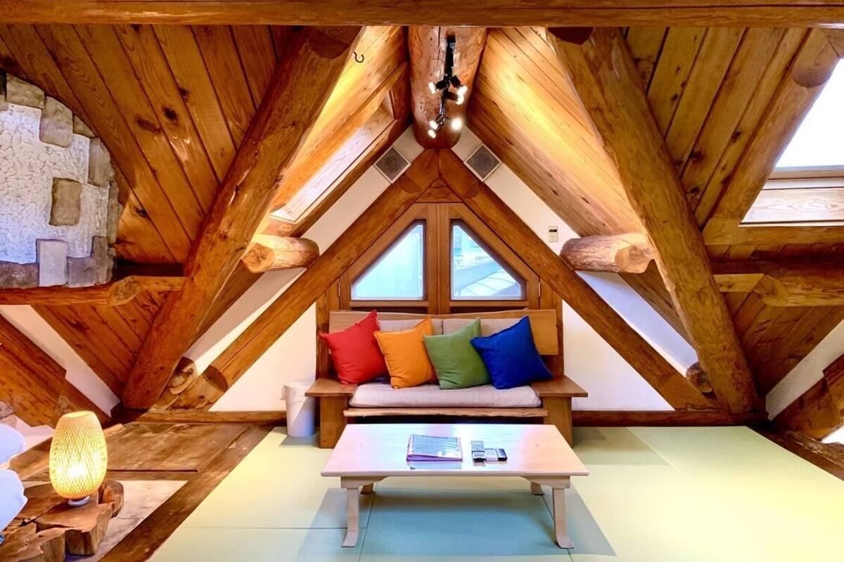 A cozy attic room with a wooden sloped ceiling, a bench with colorful cushions, and a table with books and a lamp.