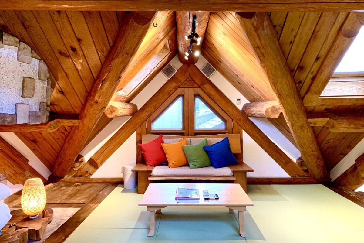 Une pièce mansardée chaleureuse avec un plafond en pente en bois, un banc agrémenté de coussins colorés et une table avec des livres et une lampe.