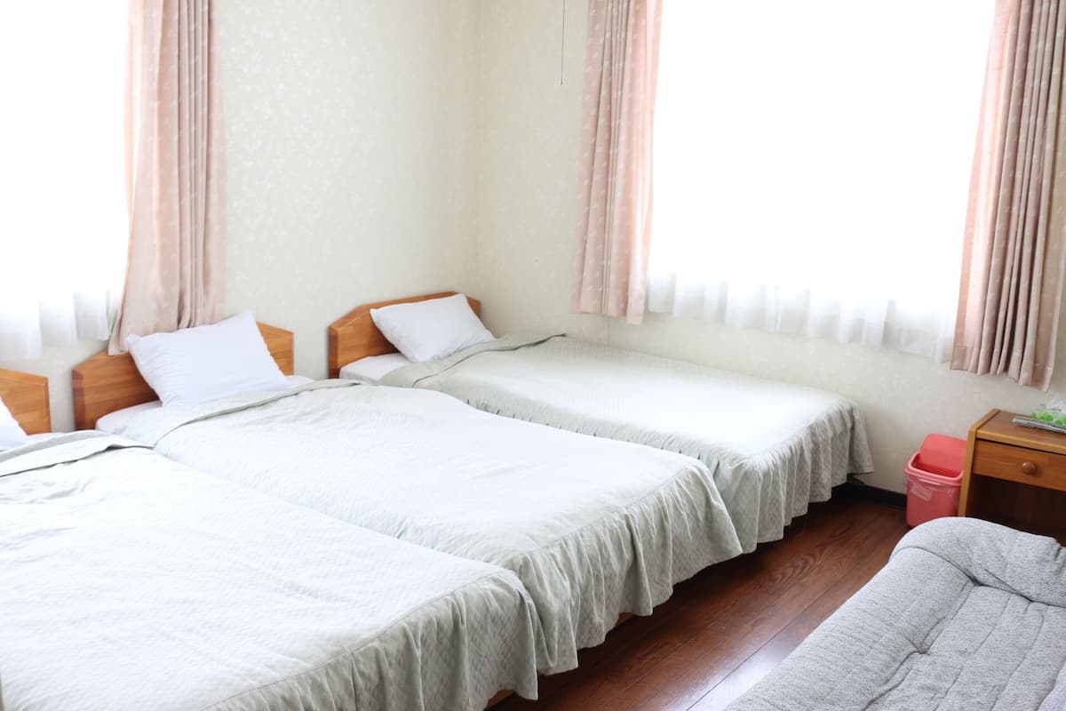 Ein Hotelzimmer mit zwei Betten, einem hölzernen Nachttisch, einer roten Mülltonne und einem Fenster mit Vorhängen.