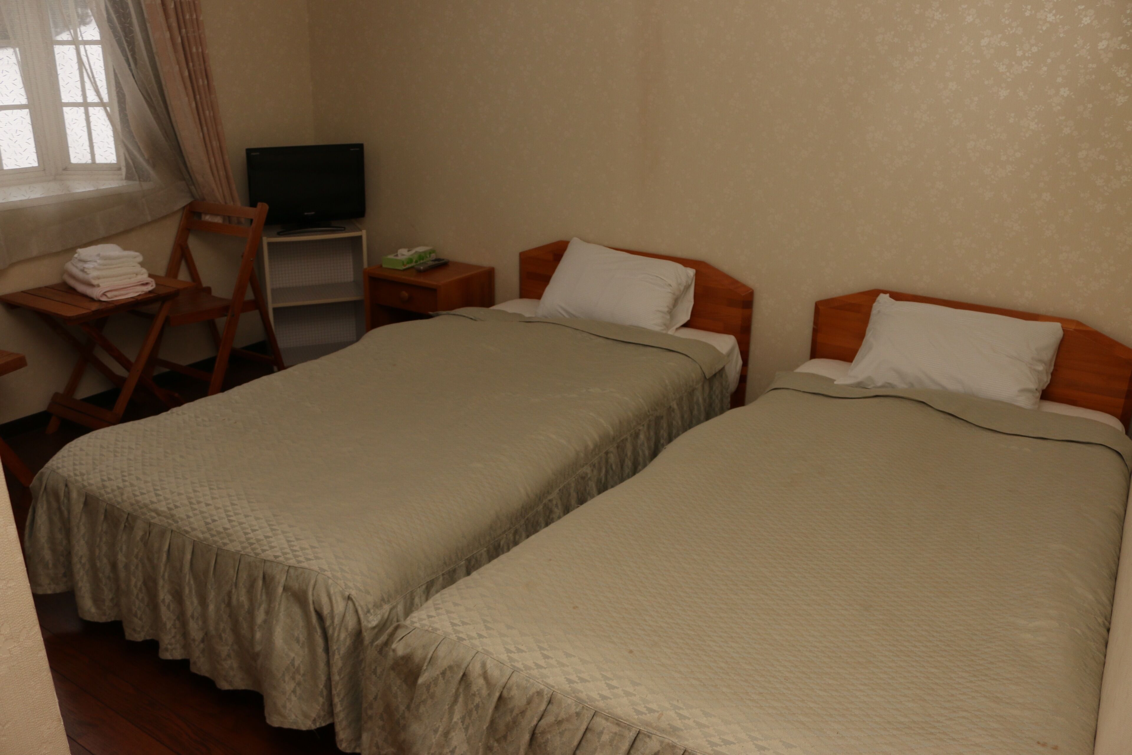 A hotel room with two single beds, a television on a stand, a chair, and a small table with a lamp.