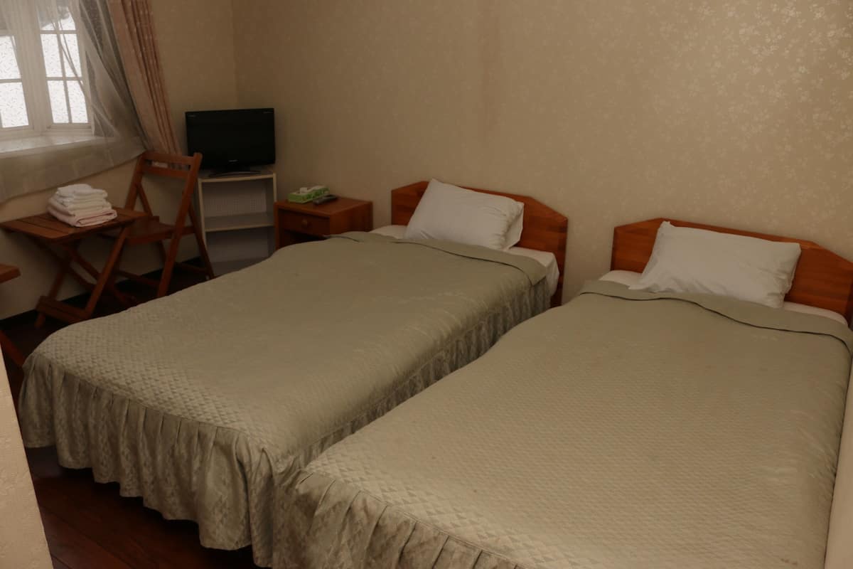 Ein Hotelzimmer mit zwei Einzelbetten, einem Fernseher auf einem Schrank, einem Sessel und einem kleinen Tisch mit einer Lampe.